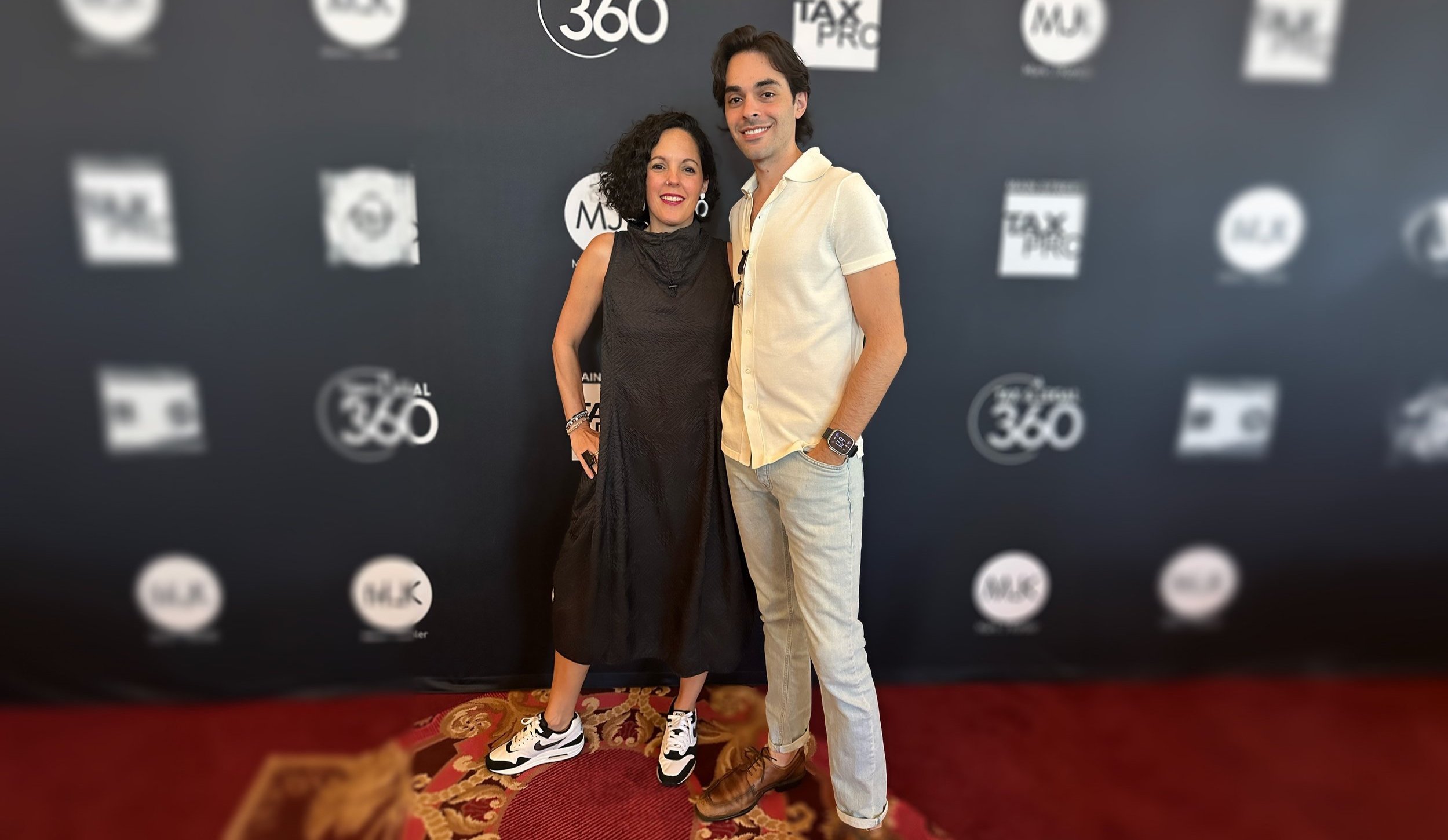 Two people standing together on a red and gold carpet at a media event, smiling, one woman in a black dress with sneakers and one man in a light-colored shirt and pants with brown shoes, against a black backdrop with various logos.